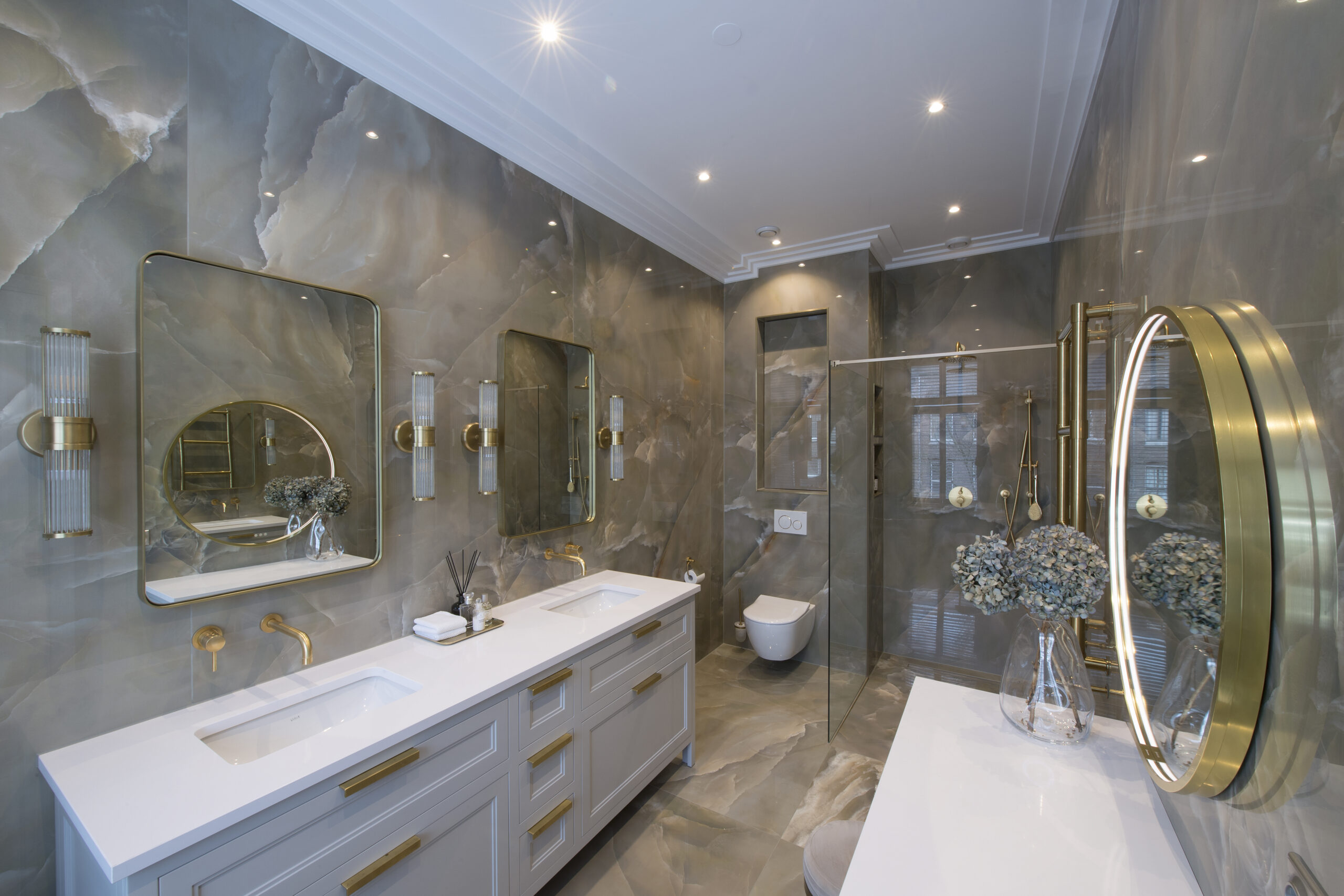Ruime luxe badkamer met marmerlook wanden, goudkleurige ronde spiegel en wastafelmeubel